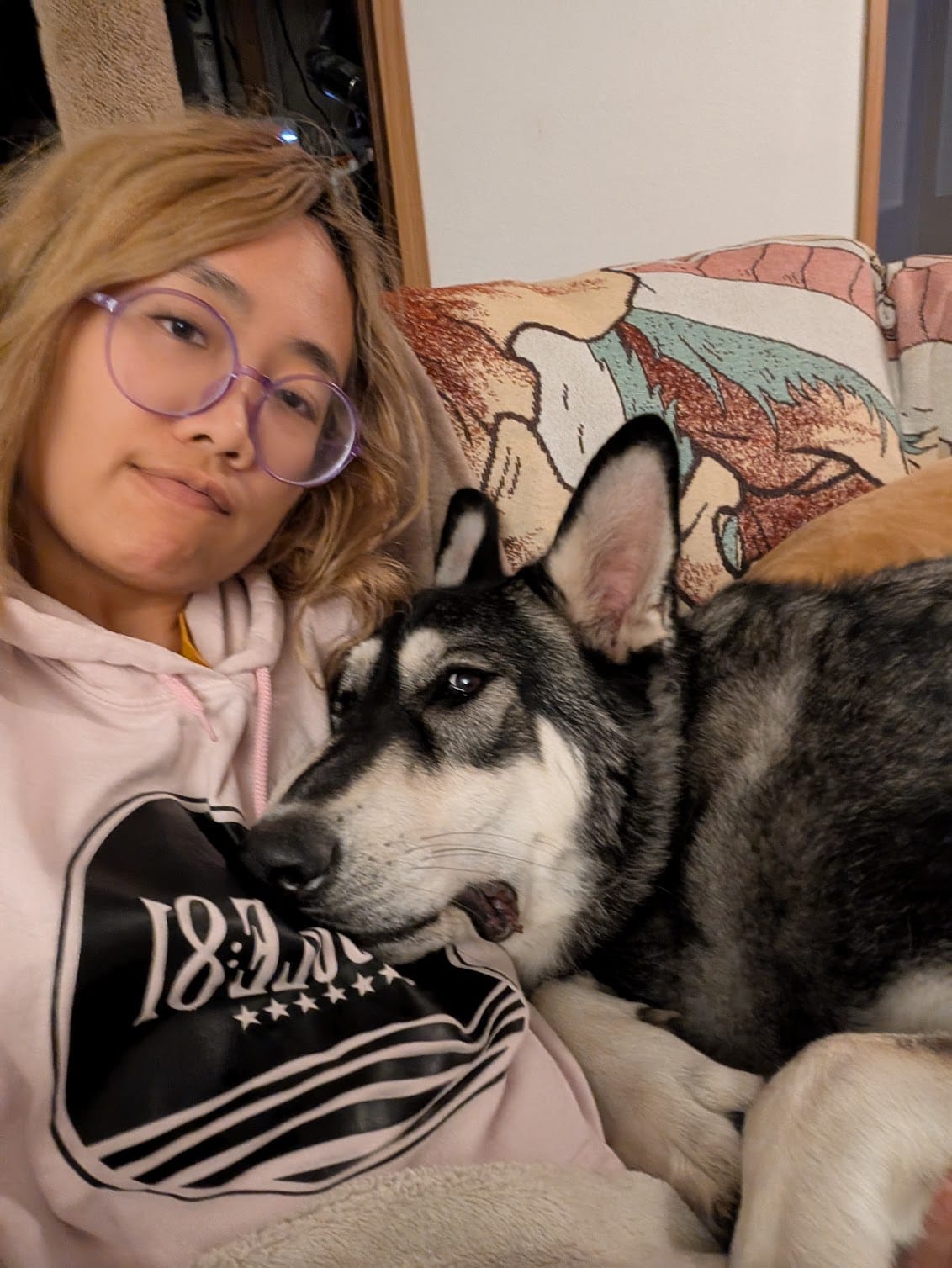 A picture of a blonde, Asian woman (me) in a pink sweatshirt, cuddling with a large black and white GSD/Akita mix dog.